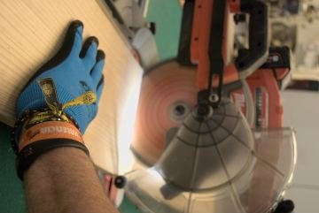 Mano con guante azul sujetando un tablero junto a la ingletadora en el taller de IRM Carpinteros en Vinaròs
